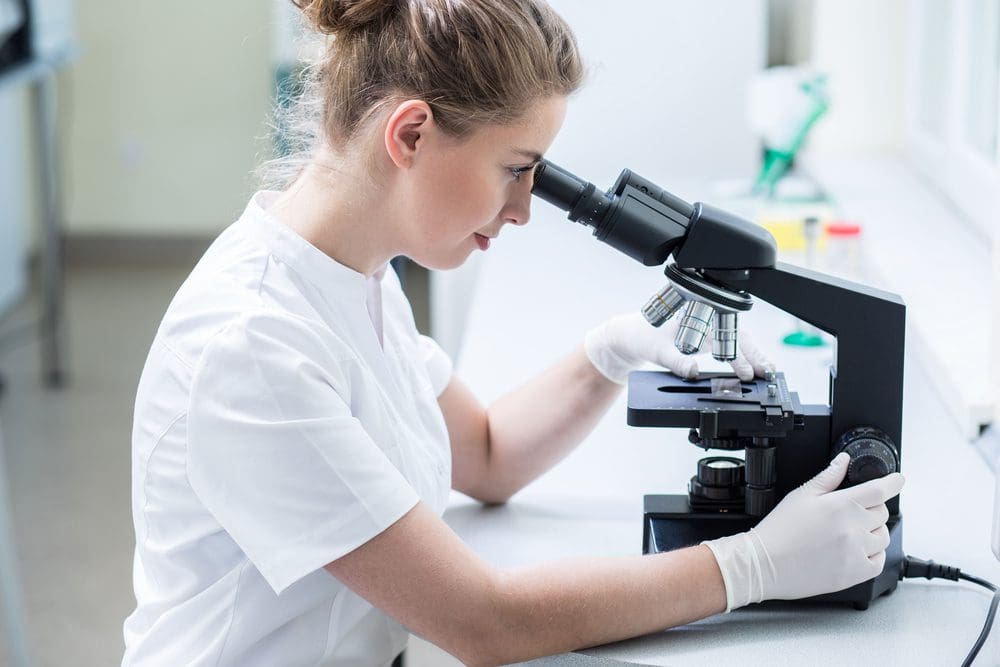 A woman in white lab coat looking through microscope.